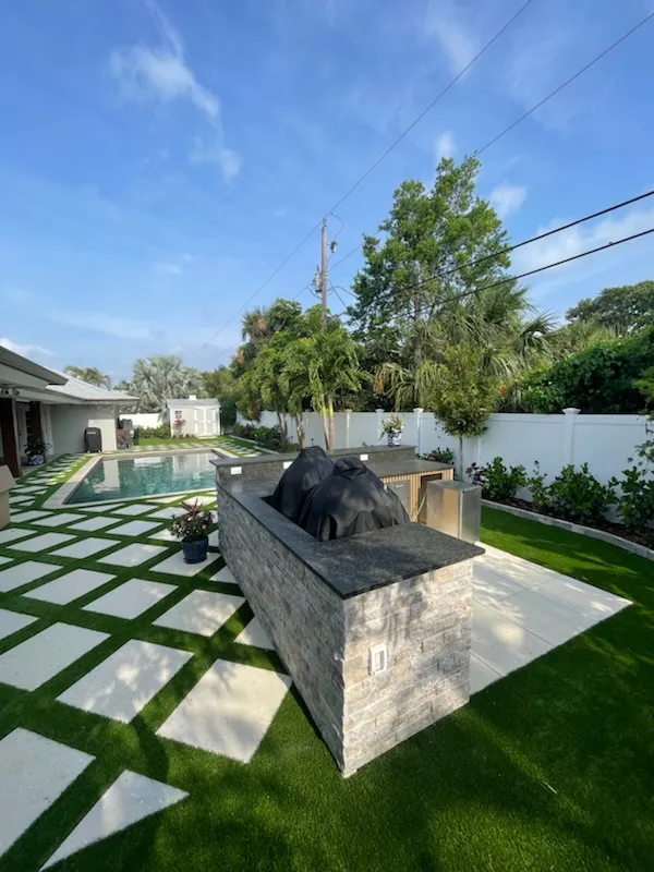 Stone grill island with granite top beside a pool and checkerboard turf-and-paver patio
